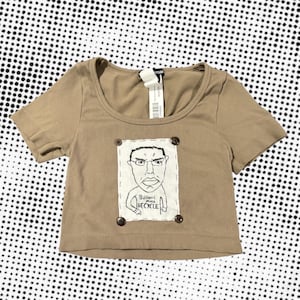 May include: A tan, ribbed crop top with short sleeves and a scoop neckline. The shirt features a hand-drawn illustration of a person with glasses and the text "Billions must RECYCLE" on the front, attached with four buttons.