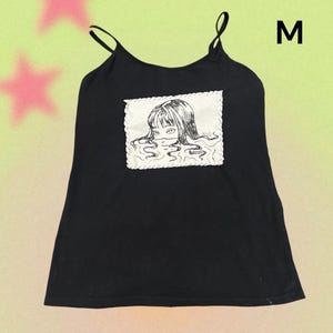 May include: Black camisole top with thin straps. A white square patch features a black and white illustration of a person's face emerging from water. The letter "M" is printed in white on the top right.