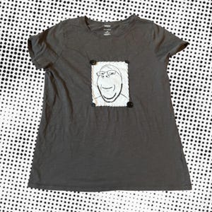 May include: Gray short-sleeve t-shirt with a cartoon face design. The design is a white rectangle with a black and white cartoon face, stitched onto the shirt with black buttons at each corner. The shirt is made of a soft material.