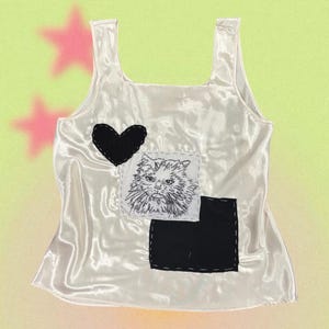 May include: A white satin vest top with a black heart and a square patch featuring an embroidered cat face. A black square patch is sewn below the cat patch. The background is a gradient of yellow and green with pink stars.