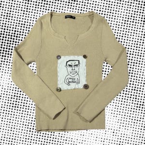 Può includere: Maglione beige a maniche lunghe con scollo tondo e piccolo scollo a V. Il maglione presenta una toppa con un disegno a cartone animato e la scritta "Billions must recycle". La toppa è fissata con quattro bottoni decorativi.