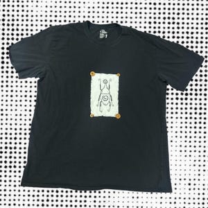 May include: Black t-shirt with a white rectangle featuring a hand-drawn insect design. The insect has a sun-like head and spiral details. The rectangle is secured with four wooden buttons.