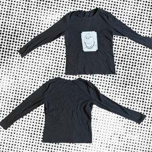 May include: Two black long-sleeved shirts. One shirt features a white square patch with a cartoon face. The other shirt is plain. The shirts are displayed on a black and white dotted background.
