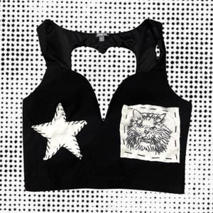 May include: Black crop top with a heart-shaped cutout and a white star and cat patch. The star and cat patches are stitched onto the top. The top is sleeveless.