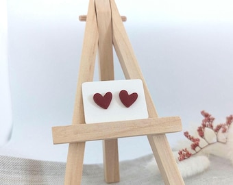 May include: A pair of burgundy heart-shaped stud earrings displayed on a white card, presented on a small wooden easel. The earrings are a deep red color, and the background is a soft white.