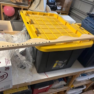 May include: A wooden measuring stick with inch markings from 1 to 12 rests on a yellow and black storage bin. The bin is on a white shelf with other items, including a box of Dr. Pepper.