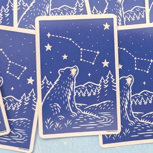 May include: A collection of blue and white cards featuring a bear looking up at a constellation. The cards have a forest and river scene at the bottom. The constellation is outlined with dotted lines and stars.