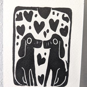 May include: Black and white linocut print featuring two dogs facing each other, with hearts above and between them. The dogs are silhouetted, with simple lines defining their features. The print is on white paper.