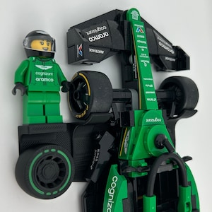 May include: A green and black Lego race car with a Lego figure in a green racing suit and helmet. The car features sponsor logos including "cognizant" and "aramco". The wheels are black with yellow detailing.