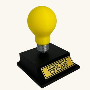 Könnte beinhalten: Eine gelbe Glühbirnen-Skulptur mit silbernem Sockel ist auf einem schwarzen rechteckigen Sockel montiert. Der Sockel hat eine goldene Plakette mit der Aufschrift "LIGHT BULB OF GLORY". Die Skulptur ist ein dekorativer Gegenstand.