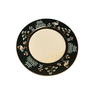 May include: A round, cream-colored plate with a dark green border. The border features a gold trim and is decorated with stylized birds, pagodas, and floral elements in shades of blue, orange, and white. A decorative plate.