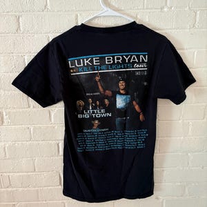 May include: Black t-shirt featuring a graphic of Luke Bryan's Kill the Lights Tour 2016. The shirt includes the tour name, the artist's name, and the names of special guests. The shirt is hanging on a white hanger.