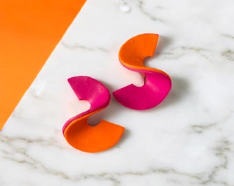 Puede incluir: Un par de pendientes abstractos con un diseño en capas. Cada pendiente tiene una forma curva, con una capa superior naranja brillante y una capa inferior rosa intenso. Los pendientes se muestran sobre una superficie de mármol blanco con un fondo naranja.