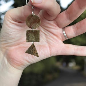 May include: A brass earring with a circular top, a square middle, and a triangular bottom. The earring is held up by a silver hook. The earring has a hammered texture and is suspended from a silver hook.