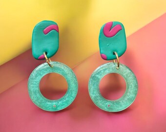 May include: A pair of handmade earrings with a teal and pink color scheme. The earrings feature a teal, rectangular top with a pink accent, connected to a translucent teal circle. The earrings hang from gold-colored hooks.
