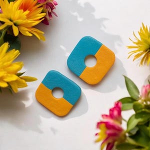 May include: Two square earrings with a central hole, each divided diagonally into blue and yellow sections. The earrings are set against a white background, surrounded by yellow and pink flowers.