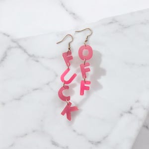 Op de afbeelding: Roze acryl oorbellen met de tekst "FUCK OFF" in individuele letterbedels. De oorbellen hebben goudkleurige oorhaken en worden gepresenteerd tegen een witte marmeren achtergrond.