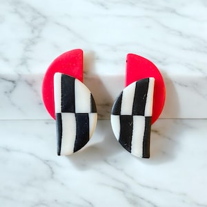 May include: A pair of statement earrings with a geometric design. The earrings are made of polymer clay and feature a black and white chequered pattern with red accents.