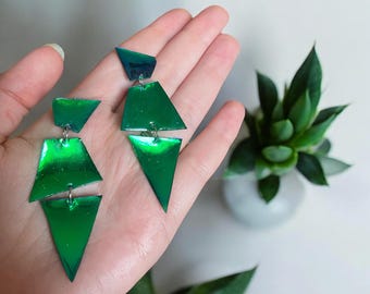 May include: Geometric green earrings with a metallic sheen. The earrings feature a layered design of angular shapes, suspended from small silver hooks. The earrings are held in a hand, with a succulent plant in the background.