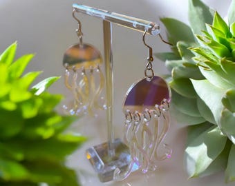 May include: Iridescent jellyfish earrings with silver-colored hooks. The earrings are displayed on a clear acrylic stand, with a succulent plant in the background. The jellyfish design has a translucent, wavy tentacle design.