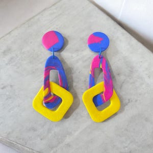 May include: A pair of vibrant, abstract earrings. Each earring features a blue and pink marbled top circle, connected to a blue and pink marbled elongated shape, and a yellow square. The earrings are set against a light gray background.