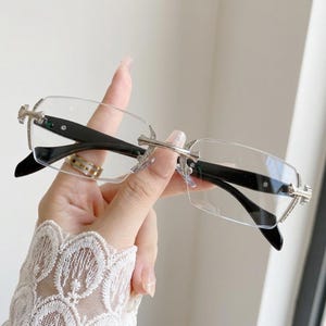 May include: Rimless rectangular spectacles featuring clear lenses and silver-toned metal hardware with intricate decorative detailing at the hinges and bridge. The temples are constructed from solid black material, providing a contrast.