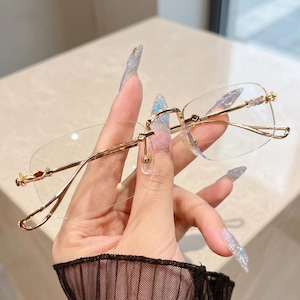 May include: Rimless eyeglasses featuring a thin, polished gold-tone metal frame with rectangular lenses. The temples and bridge exhibit intricate, textured detailing, and the nose pads are clear, adjustable silicone components.