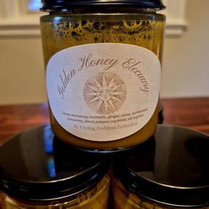 May include: A stack of glass jars filled with golden honey. Each jar has a black lid and a label that reads "Golden Honey Elecuary" with a sun design. The label also lists ingredients like turmeric and ginger.