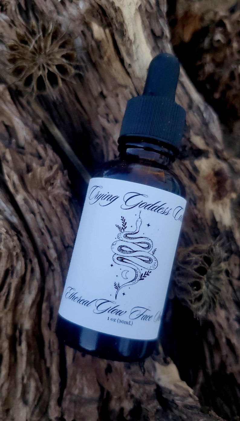 Op de afbeelding: Een glazen fles met een zwarte druppelaar en een wit etiket. Het etiket heeft een zwart-witte illustratie van een slang met een halve maan en sterren. De tekst op het etiket luidt "Trying Goddess Co. Ethereal Glow Face Serum 1 oz (30ml)"