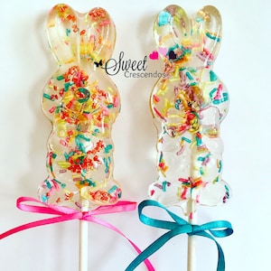 May include: Two clear bunny-shaped lollipops with colorful sprinkles, tied with pink and blue ribbons.