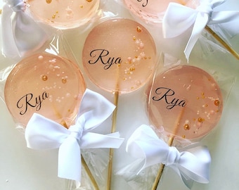 May include: Five pink and white lollipops with the name "Rya" written on each one. The lollipops are wrapped in clear cellophane and tied with white bows.