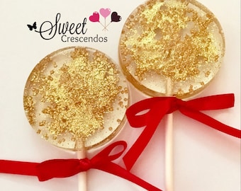 May include: Two round lollipops with gold glitter on a clear base. Each lollipop is tied with a red ribbon and attached to a white stick. The lollipops are on a white background. The text "Sweet Crescendos" is visible in the top left corner of the image.