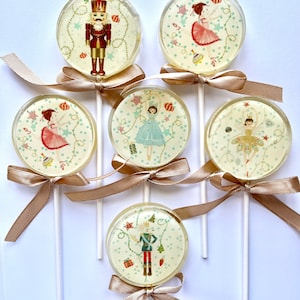 May include: Six round lollipops with festive designs. The lollipops feature images of a nutcracker, a ballerina, and a fairy. The lollipops are on white sticks with gold ribbon bows.