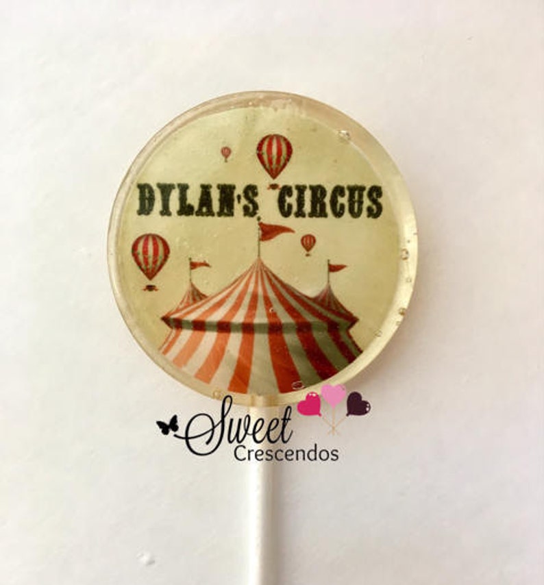 Może przedstawiać: Okrągły lizak z białym tłem i kolorowym wzorem namiotu cyrkowego. Tekst "Dylan's Circus" jest nadrukowany czarnymi literami na lizaku.