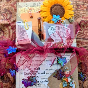 May include: A decorative paper packet featuring a yellow felt sunflower, a sticker reading Emotionally Attached To Fictional Characters, a scarecrow cutout, and various paper scraps with text. The bundle is tied with a sheer dark red ribbon.