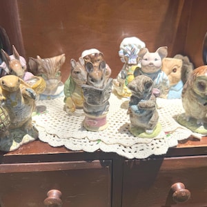 May include: A collection of ten small ceramic animal figurines featuring characters in various poses, including a turtle with a basket, a rabbit reading a newspaper, and an owl, all arranged on a white crocheted doily atop a wooden cabinet.