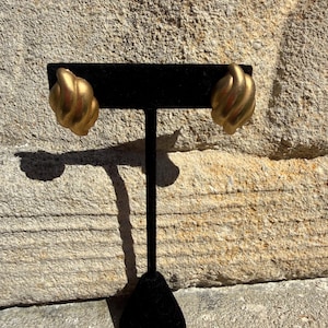 May include: A pair of gold-colored, textured clip-on earrings. Each earring has a rounded, swirled design. The earrings are displayed on a black velvet earring stand against a textured stone background.