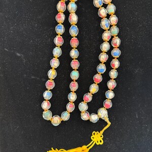 May include: A beaded necklace with clear, round beads containing colorful images. The beads are strung on a gold-colored chain, and the necklace has a yellow tassel. The beads have a variety of colors, including red, blue, and green.