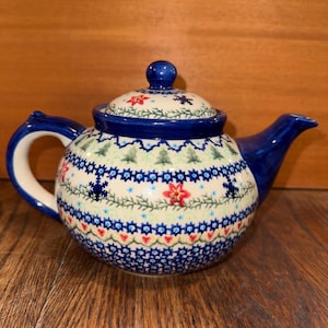 May include: A ceramic teapot featuring a rounded body and a matching lid. The surface is decorated with a festive pattern of green pine trees, red poinsettia flowers, and blue snowflake motifs arranged in horizontal bands on a cream background.