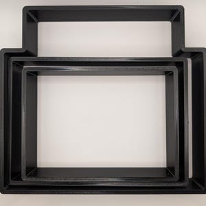May include: Three black, rectangular cookie cutters of varying sizes are nested together. The largest cutter has a handle-like extension on top. The cutters are made of a matte black material and are set against a white background.
