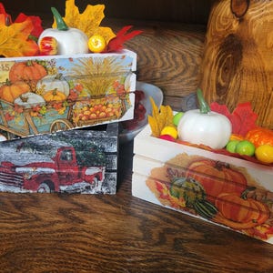 May include: Two white wooden crates decorated with fall-themed images. The top crate has a red truck and the bottom crate has a pumpkin and fall leaves. Both crates have a white pumpkin and fall leaves on top.