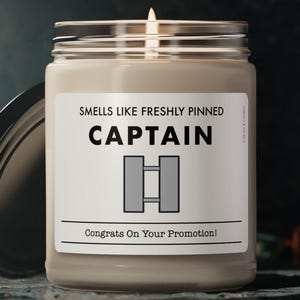 May include: A lit candle in a glass jar with a black lid. The label reads "Smells Like Freshly Pinned CAPTAIN" with a rank insignia and "Congrats On Your Promotion!" in white text. The candle sits on a dark, textured surface.