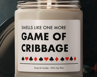 May include: A beige candle in a glass jar with a black lid. The label reads "Smells like one more Game of Cribbage". The label also has a row of playing card suit symbols.