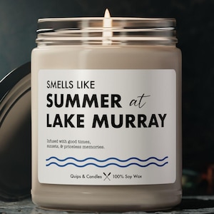 May include: A lit candle in a glass jar with a silver lid. The label reads "SMELLS LIKE SUMMER at LAKE MURRAY." The candle is on a dark surface. The text also says "infused with good times, moments, & priceless memories."