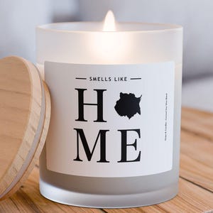 Peut inclure: Une bougie en verre givré avec un couvercle en bois. La bougie est allumée et l'étiquette indique "Smells Like Home".