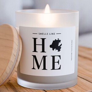 May include: A white frosted glass candle with a wooden lid. The candle has a black label that says "Smells Like Home" with a small silhouette of a house.