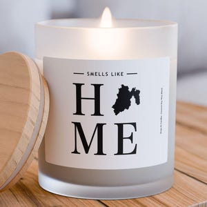 Peut inclure: Une bougie en verre givré blanc avec une étiquette noire et blanche qui indique "Smells Like Home". La bougie est allumée et possède un couvercle en bois.