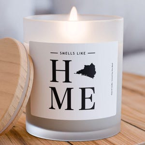 May include: A white frosted glass candle with a wooden lid. The candle has a black label that says "Smells Like Home" with a silhouette of a state shape. The candle is lit and sitting on a wooden table.