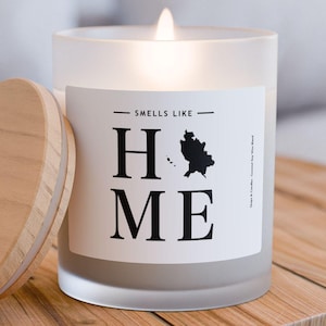 Könnte beinhalten: Eine gefrostete Glaskerze mit einem weißen Etikett, auf dem in schwarzer Schrift "Smells Like Home" steht. Die Kerze ist angezündet und hat einen Holzdeckel.