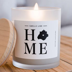 May include: A frosted glass candle with a wooden lid. The candle is lit and the label reads "Smells Like Home".